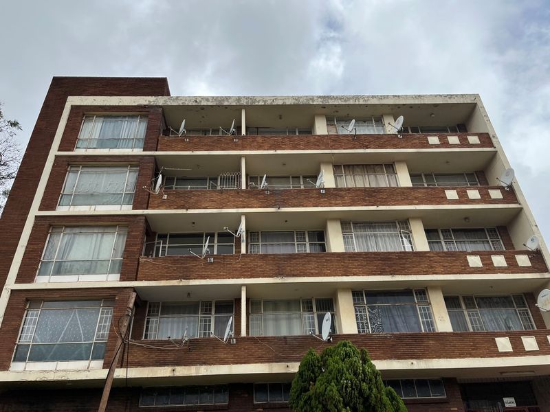 Multi-story residential building facade featuring exposed brickwork, multiple balconies, and metal-framed windows with various satellite dish installations.