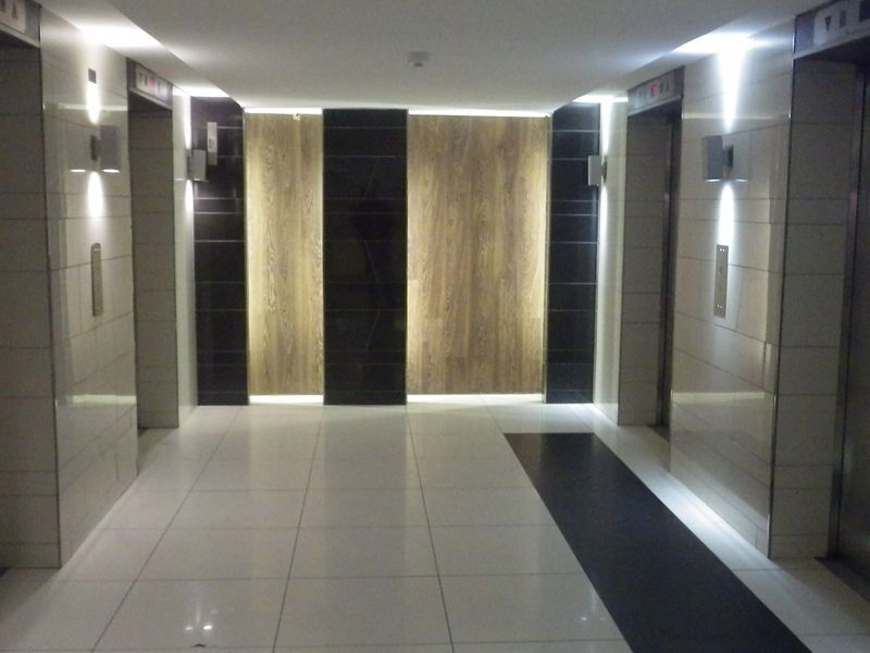 A modern, well-maintained apartment building hallway featuring tiled flooring, recessed lighting, and multiple elevator access points with contemporary wall finishes.
