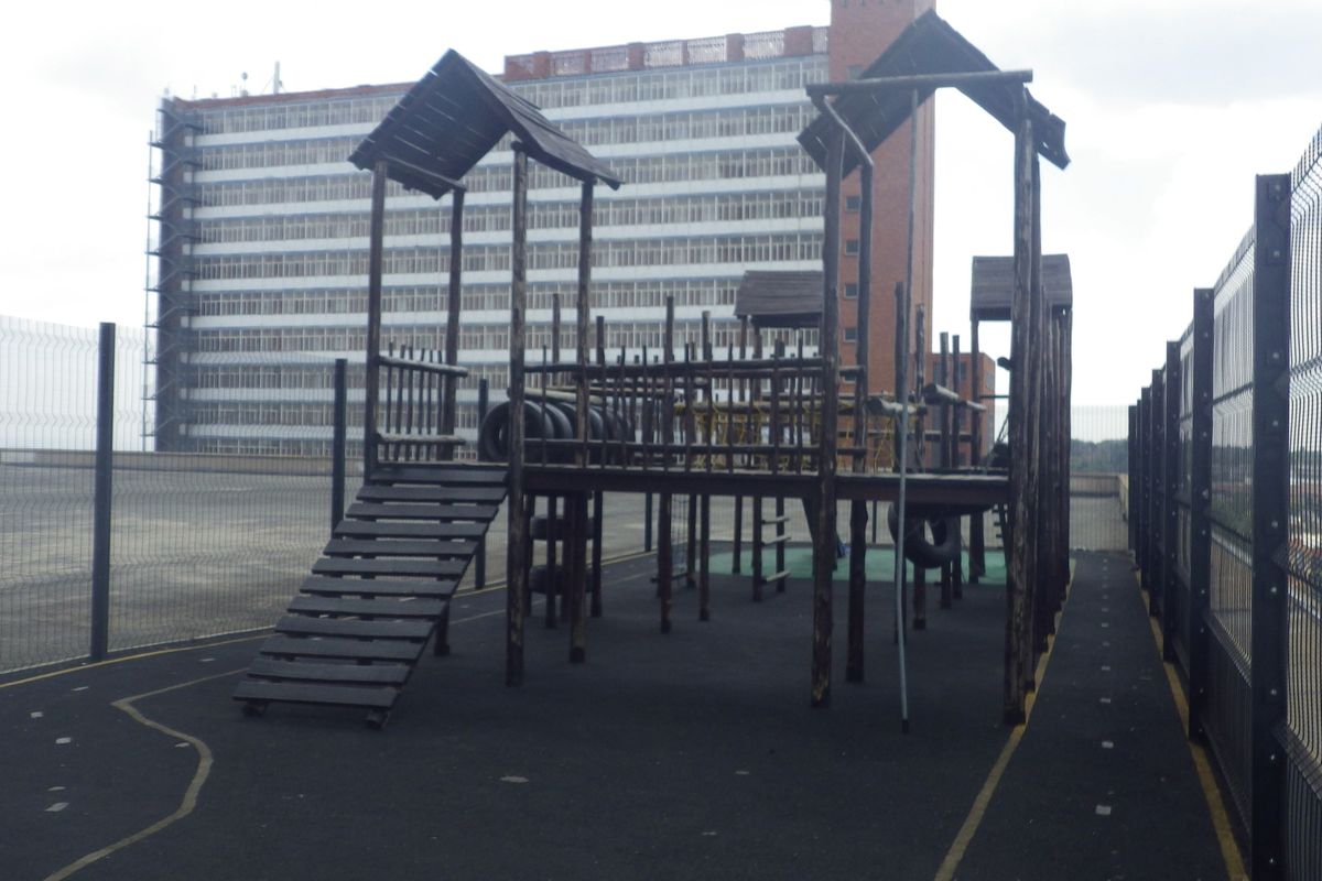 Outdoor communal play area featuring a timber climbing structure set on a paved surface, enclosed by security fencing.
