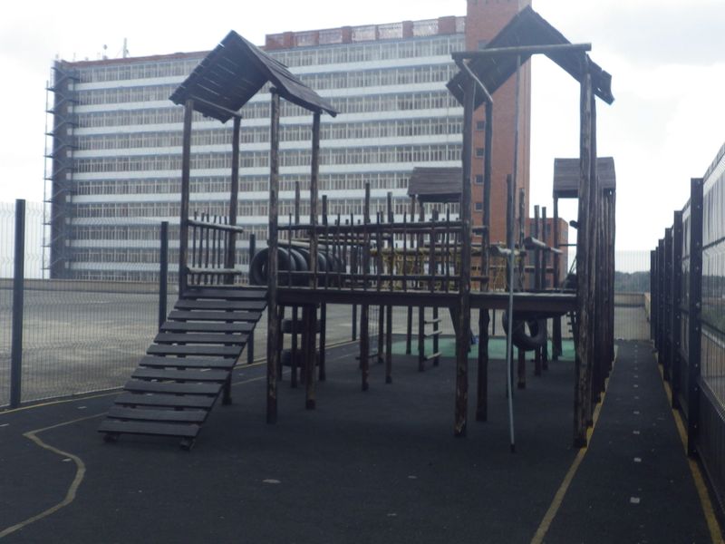 Outdoor communal playground area featuring timber climbing structures and security fencing on a paved surface.