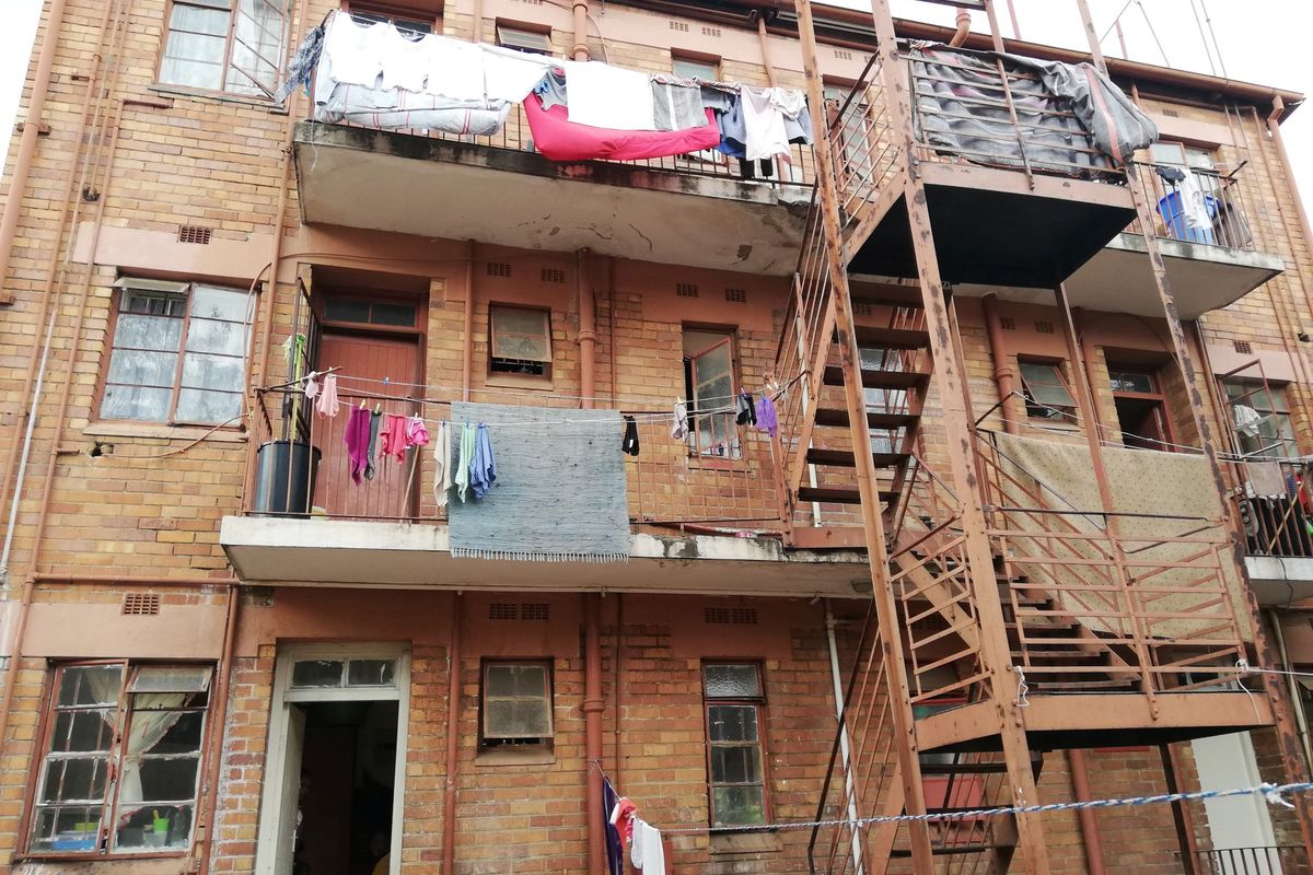 Rear elevation of a multi-story brick apartment building featuring external steel staircases and concrete balconies showing signs of significant wear and weathering.