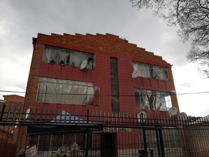 Front elevation of a multi-story brick apartment building featuring a stepped parapet design, large windows, and perimeter security fencing.