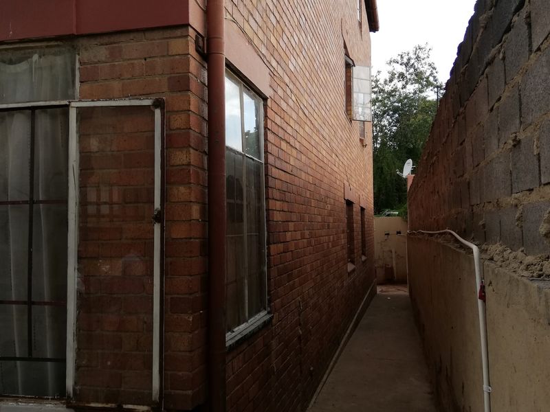 A narrow side passage between the brick building facade and a boundary wall, featuring a concrete walkway and external drainage infrastructure.