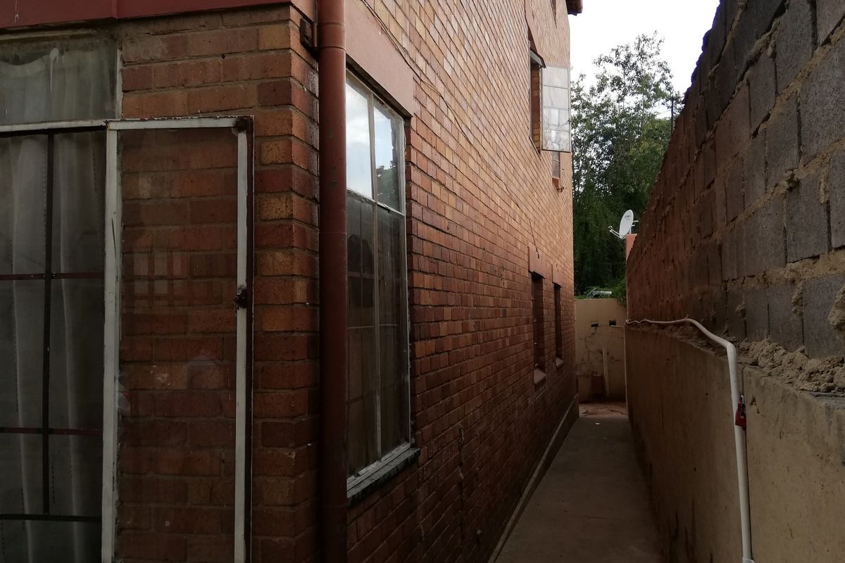 A narrow side passage between the brick building facade and a boundary wall, featuring a concrete walkway and external drainage infrastructure.