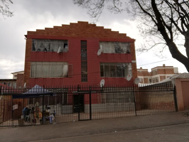 Front elevation of a multi-story residential flat building featuring a red facade, stepped brick parapet, and perimeter security fencing.